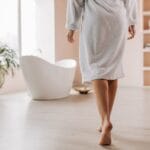 Osoba w szlafroku idąca do wanny w jasnej łazience z drewnianą podłogą